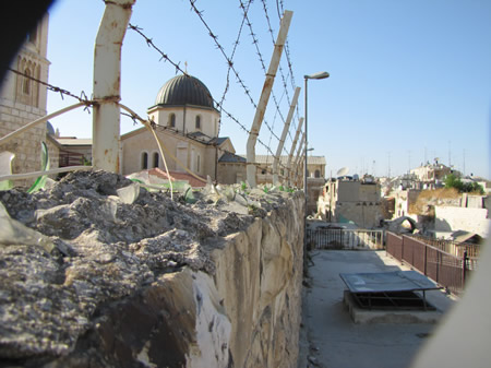 A stone wall in Jerusalem with barbed wire and glass embedded into concrete forms a barrier.