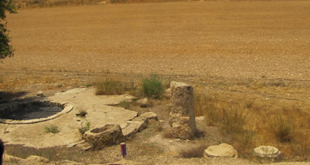 A Roman mile marker still stands beside a cistern and a water trough along a modern road in Israel. 